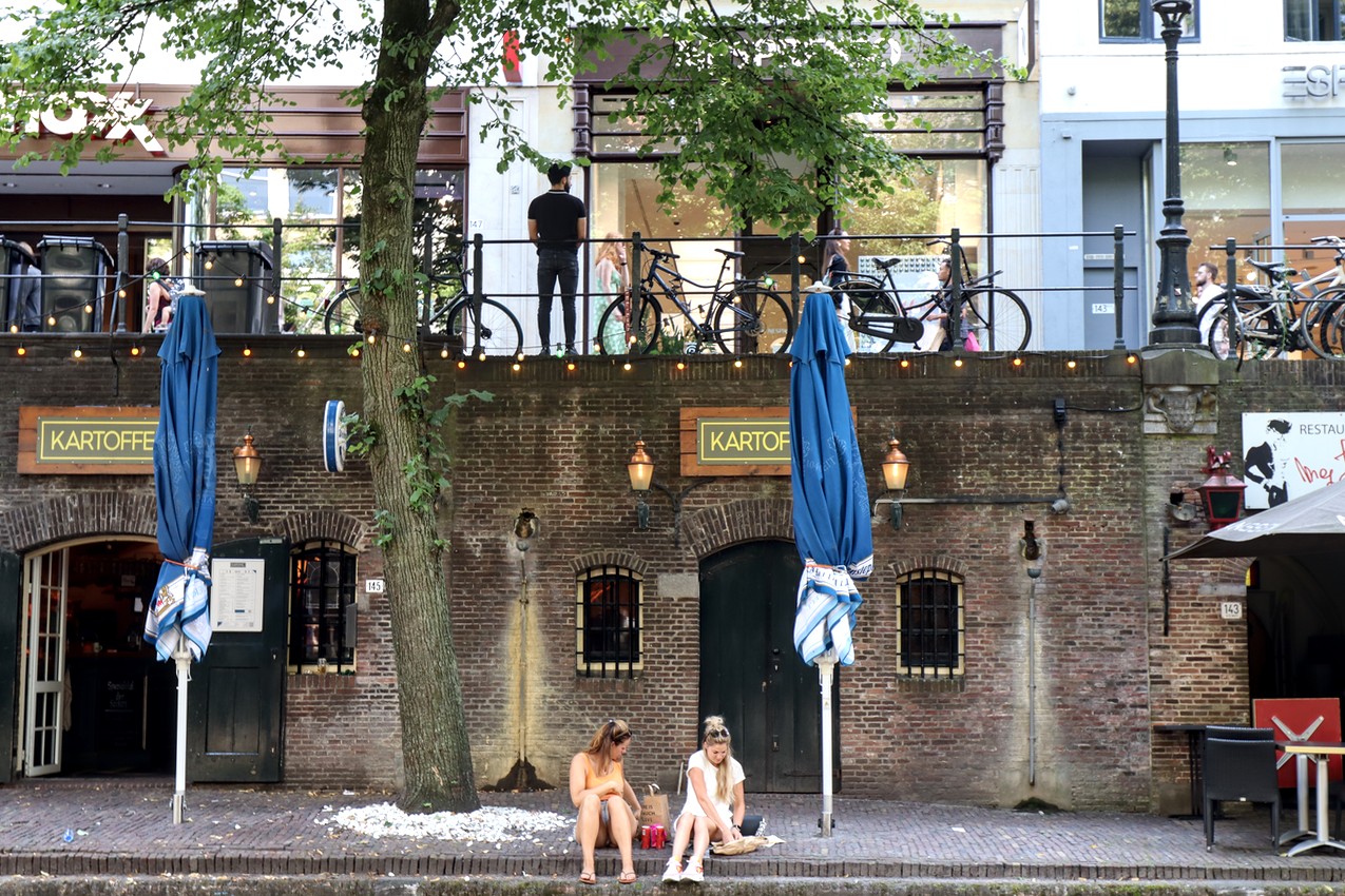 Two women eat lunch along a canal; on the second level above them a man leans on a railing