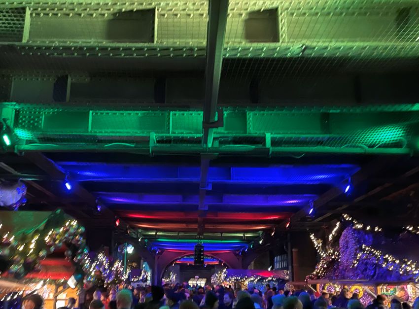 Rainbow lights under a train bridge, with Christmas market huts lining the sides and a large crowd packing the area